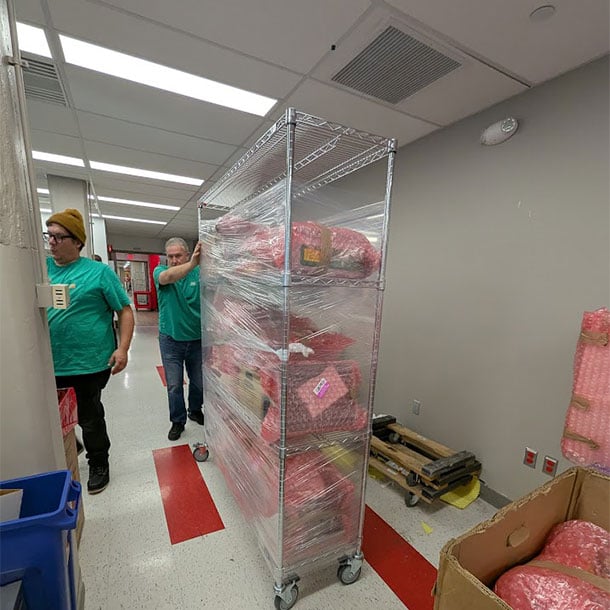 Lab Movers workers transporting a plastic-wrapped wire shelving cart loaded with supplies through a research facility corridor