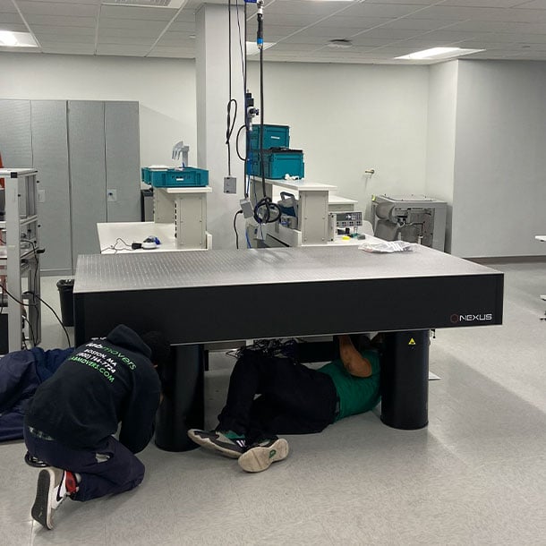 Lab Movers technicians crawling beneath a large Nexus optical table to level and adjust pneumatic isolation legs during laboratory setup