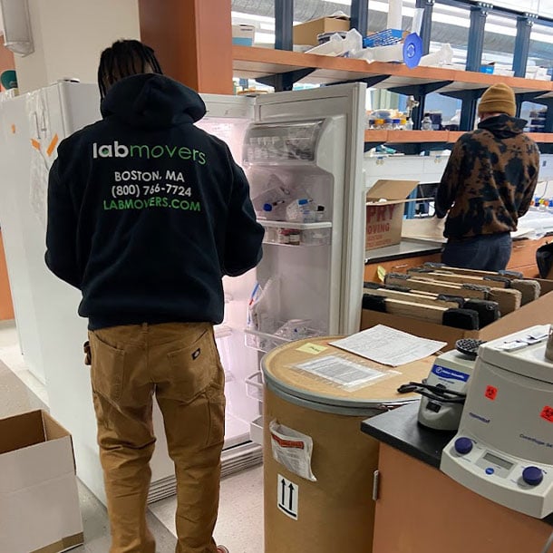 Lab Movers crew member in branded hoodie emptying and preparing a laboratory refrigerator for relocation while colleagues pack nearby equipment
