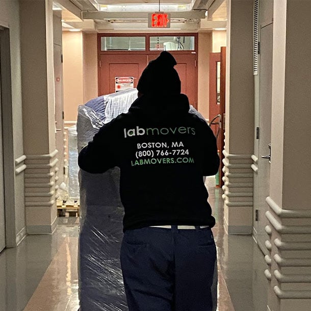 Lab Movers crew member in branded hoodie transporting a plastic-wrapped piece of laboratory equipment through a research building corridor